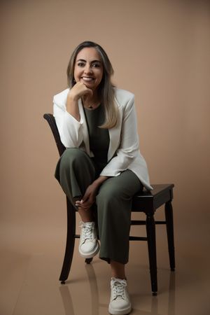 Professional fashion portrait series showing someone seated on a dark chair wearing a white blazer and dark pants.