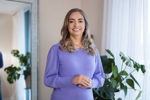 Woman in purple sweater smiling and posing in bright room with houseplants and white curtains.