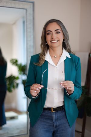 Professional wearing a teal blazer and white blouse poses in a bright modern office space with a window view.