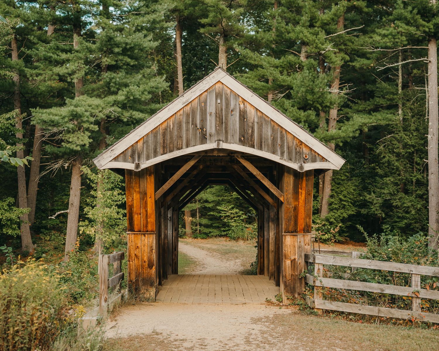 A rustic wooden covered bridge with brown timber frame leads into a green forest along a dirt path.