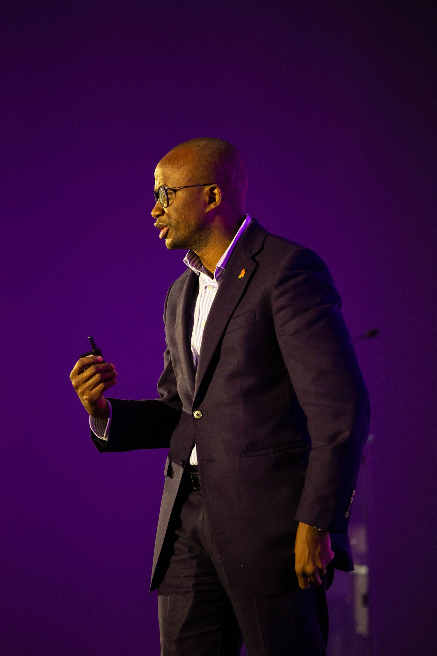 A speaker in a dark suit gestures while presenting on stage with dramatic purple lighting in the background.