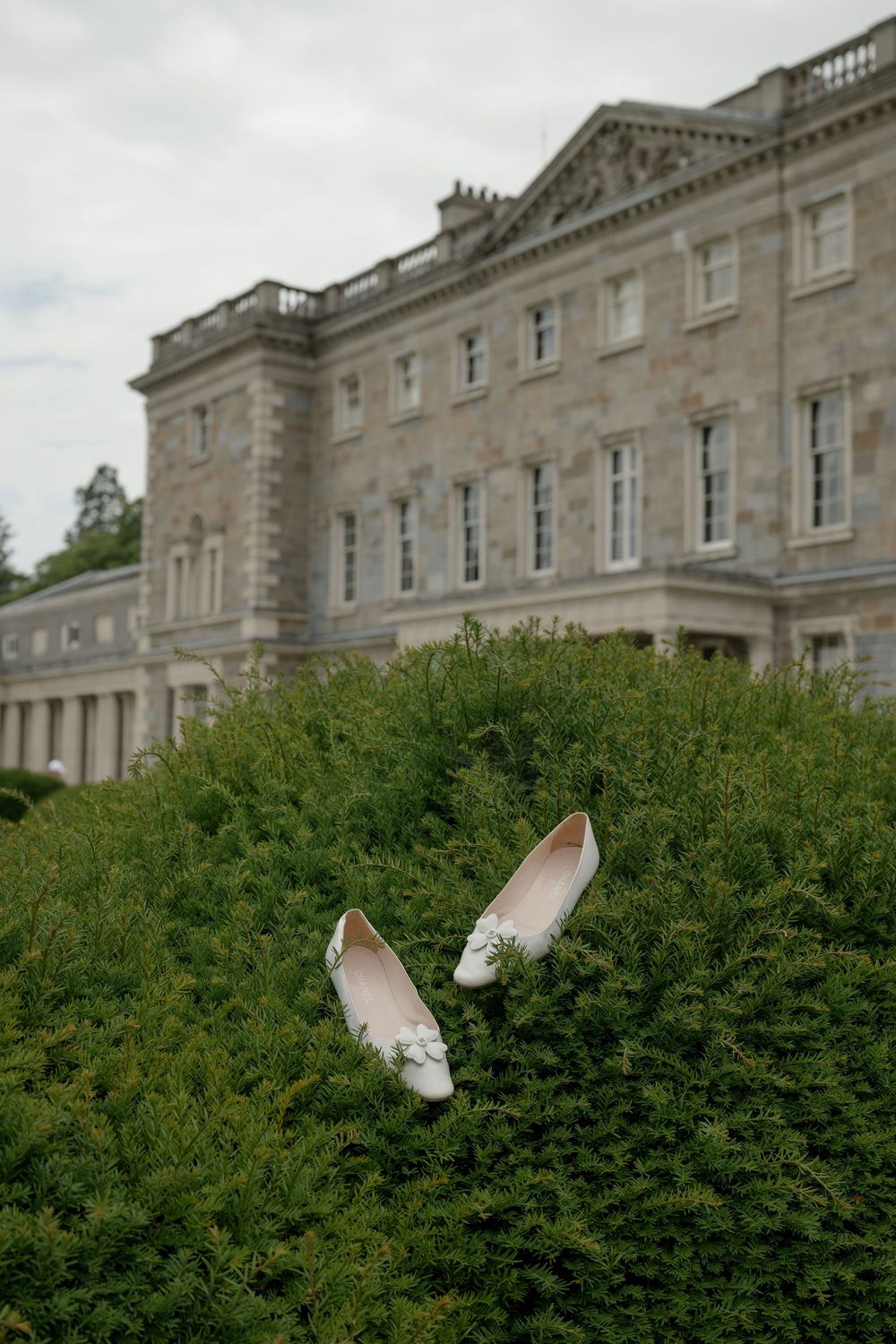 White wedding shoes displayed artistically in front of a grand stone manor house with manicured hedges.