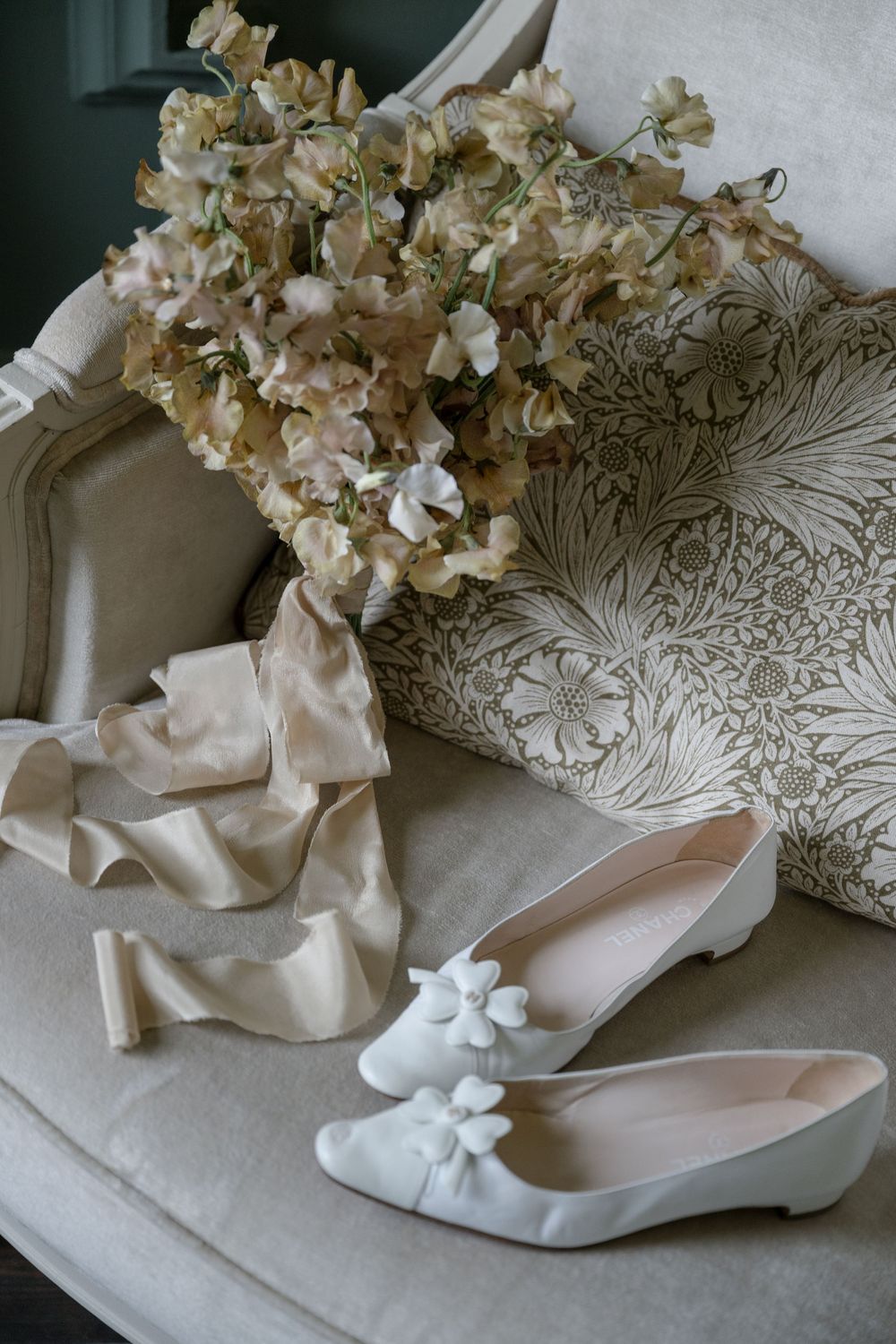 Delicate white bridal ballet flats and dried hydrangea flowers arranged on textured linen fabric.