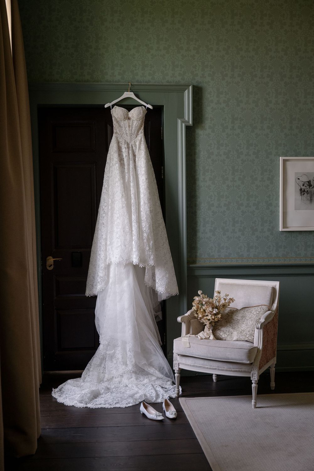 An elegant white lace wedding dress hangs in a classic room with a vintage chair and soft lighting.