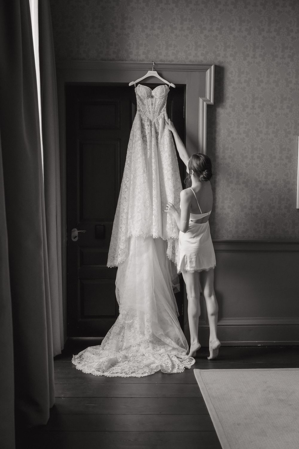 Elegant white lace wedding dress hanging in a room with dramatic lighting and shadows.