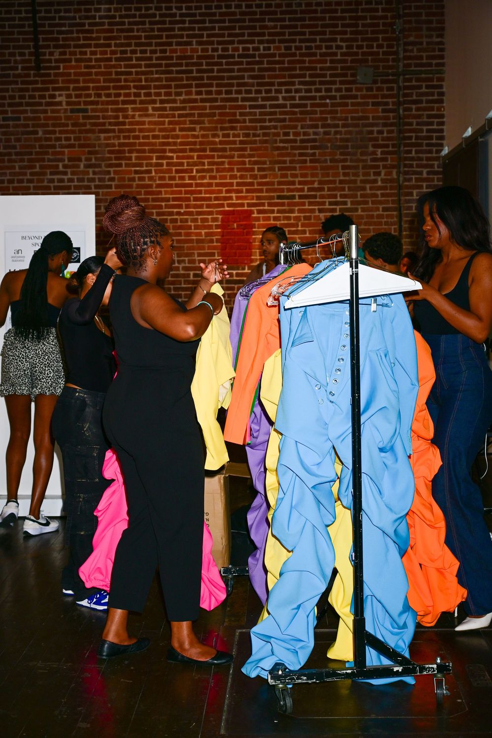 People browse through colorful clothing on a rack against exposed brick wall at what appears to be a fashion event or sale.
