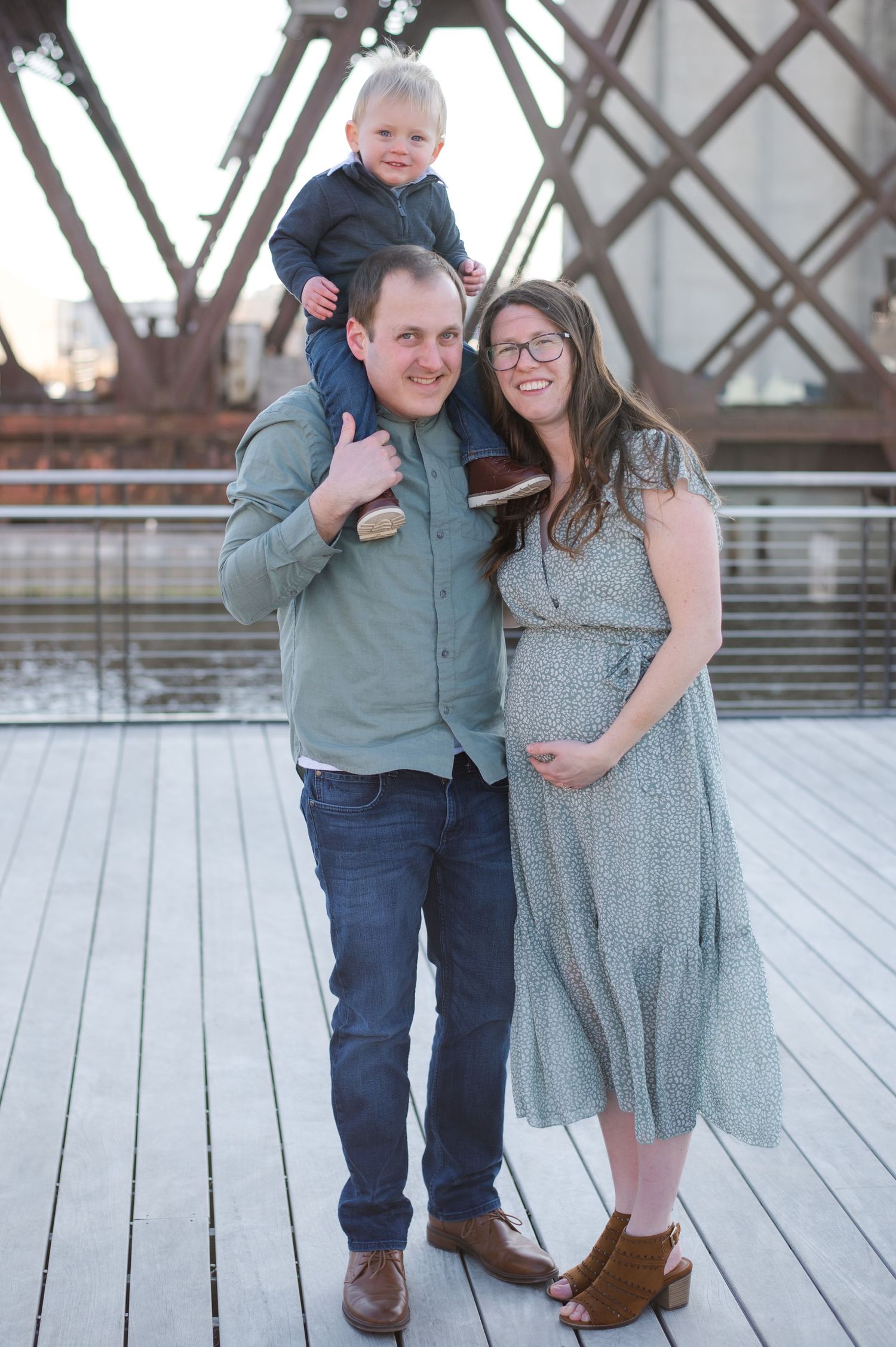 A family of three stands lovingly together on a wooden deck in coordinating sage green and grey outfits.