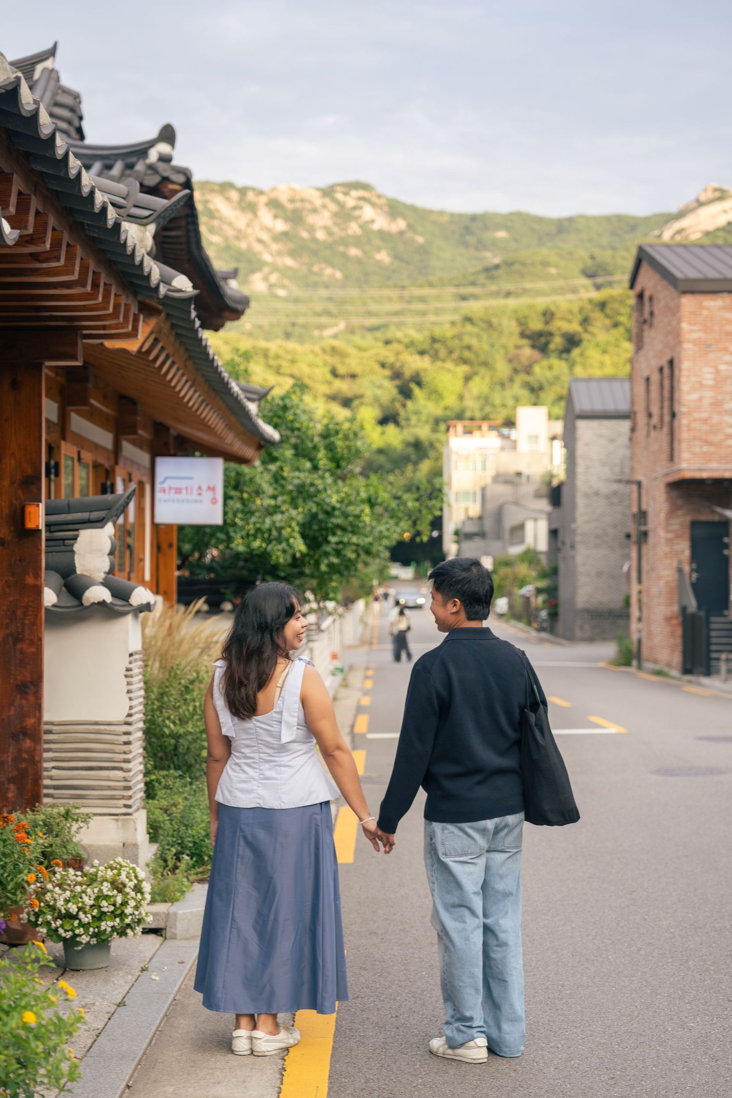Two people walking down a scenic path lined with traditional Korean architecture and mountain landscapes.