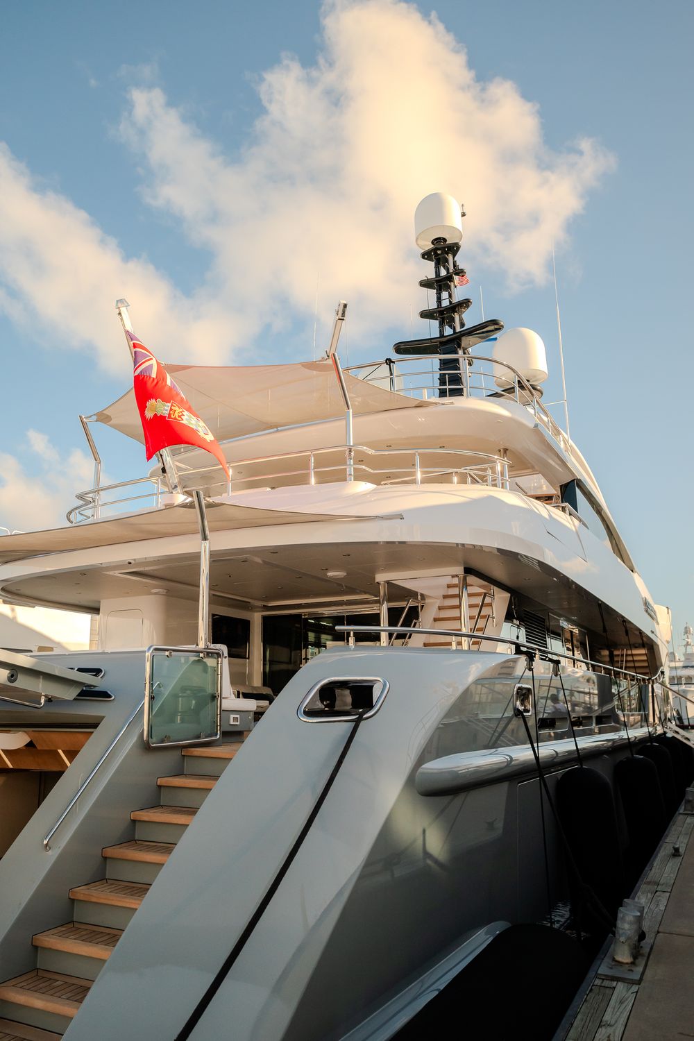 Luxury yacht with multiple decks and communication equipment displays a red flag against blue sky with white clouds.