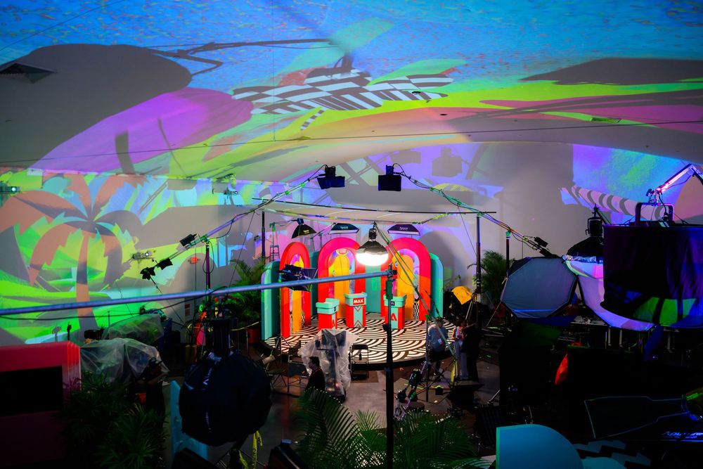 Concert stage setup with colorful psychedelic light projections and musical equipment silhouetted against vibrant backdrop.