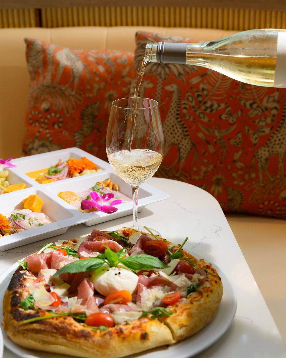 White wine being poured next to a gourmet pizza and appetizer platter on a table with red patterned cushions.
