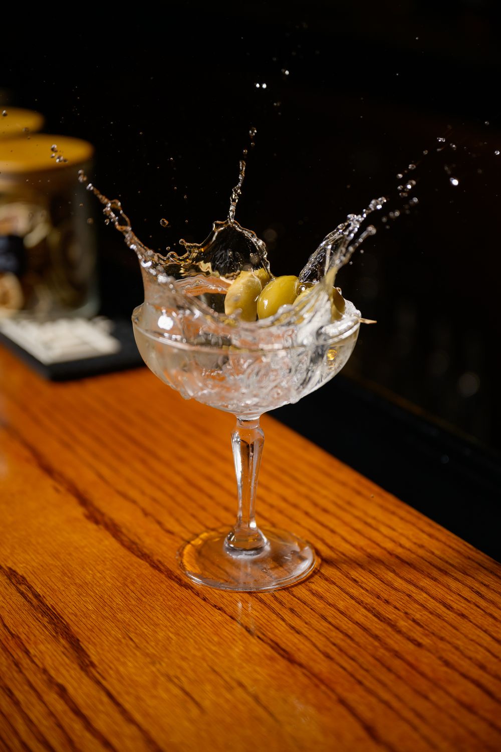 Cocktail splashing in a coupe glass with olives on a wooden bar counter creating dynamic liquid motion.