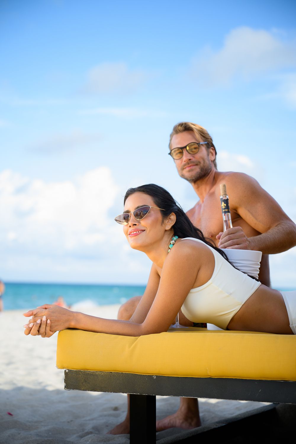 Couple relaxing on a yellow lounge chair at a sunny beach with turquoise water.