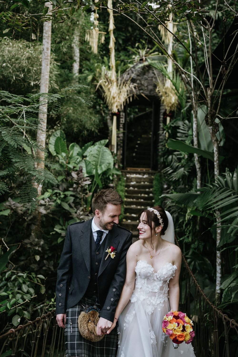 A wedding couple shares a joyful moment on a wooden walkway surrounded by tropical foliage.