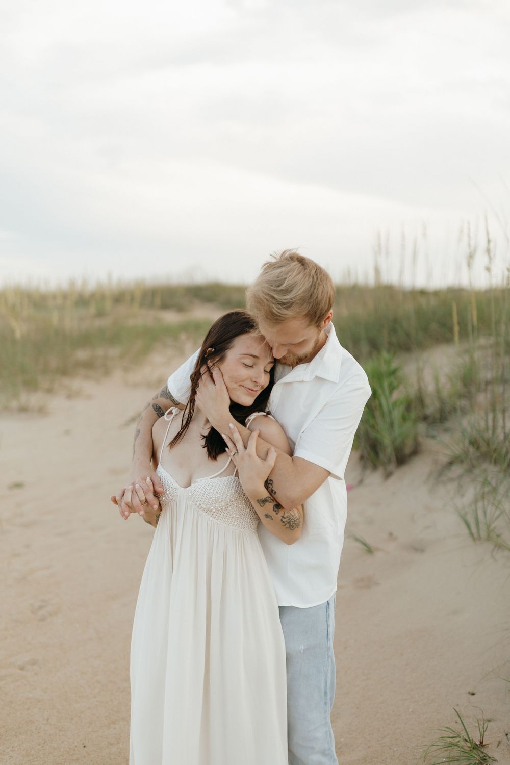A couple shares a tender embrace on a beach with sand dunes and sea grass in the background.