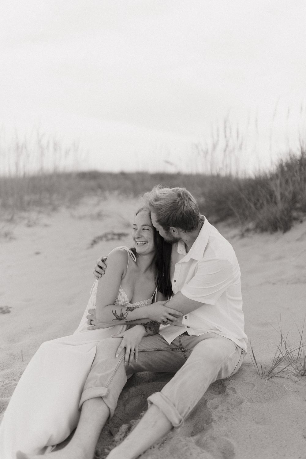 A romantic black and white photo of a couple embracing on a sandy beach with tall grass in the background.
