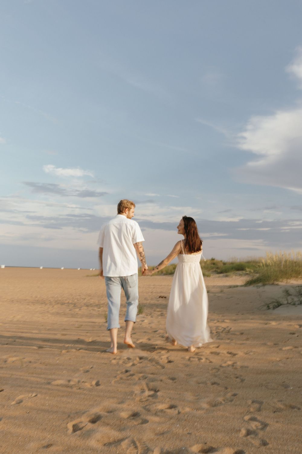 A couple walks hand in hand on a sandy beach at sunset with footprints trailing behind them in the sand.