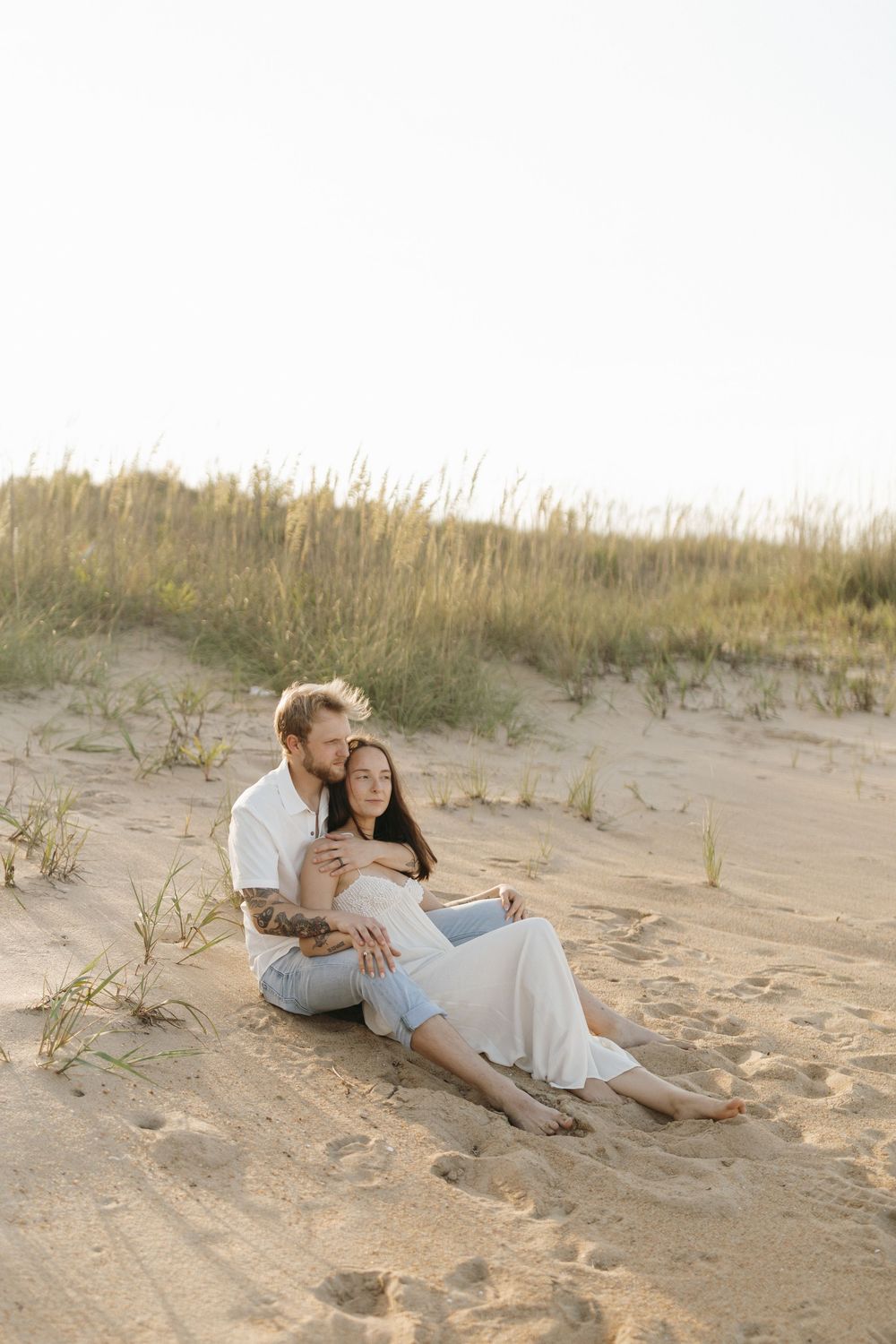 A couple shares a romantic moment lying on a sandy beach with tall grass in the background during golden hour.