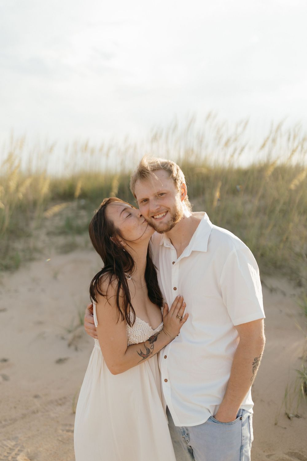 A loving couple shares an intimate moment on a sandy beach path with tall grass swaying in the background.