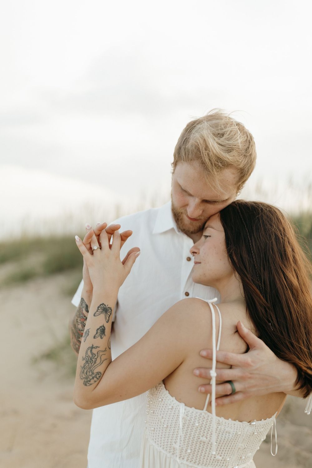 A romantic beach photoshoot shows a couple in white embracing intimately on sandy dunes at sunset.