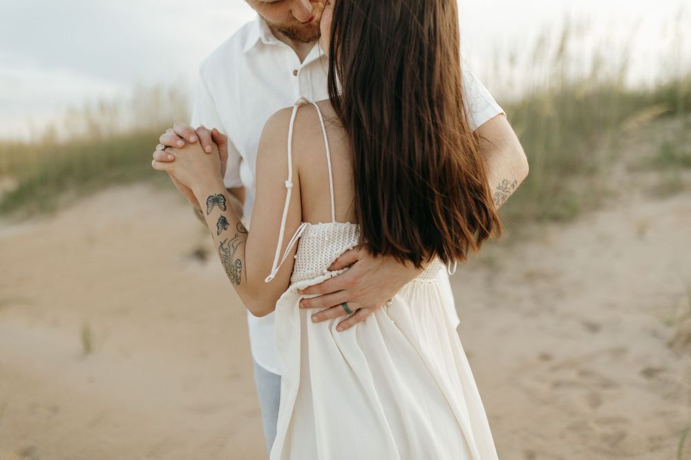 Romantic couple in white outfits dancing together on sandy beach dunes at sunset.