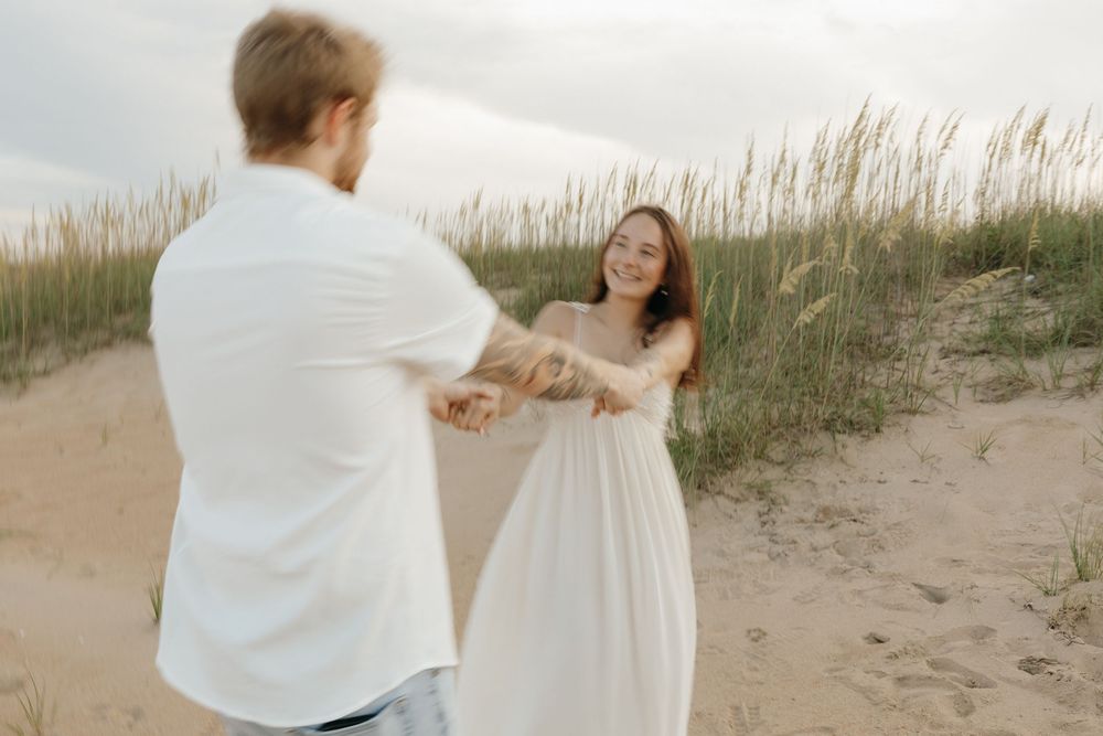 A couple in white outfits dances joyfully on a beach with dune grass swaying in the background.