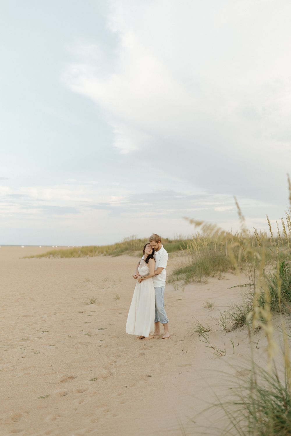 A couple embraces on a serene beach with sand dunes and sea grass swaying in the soft coastal breeze.