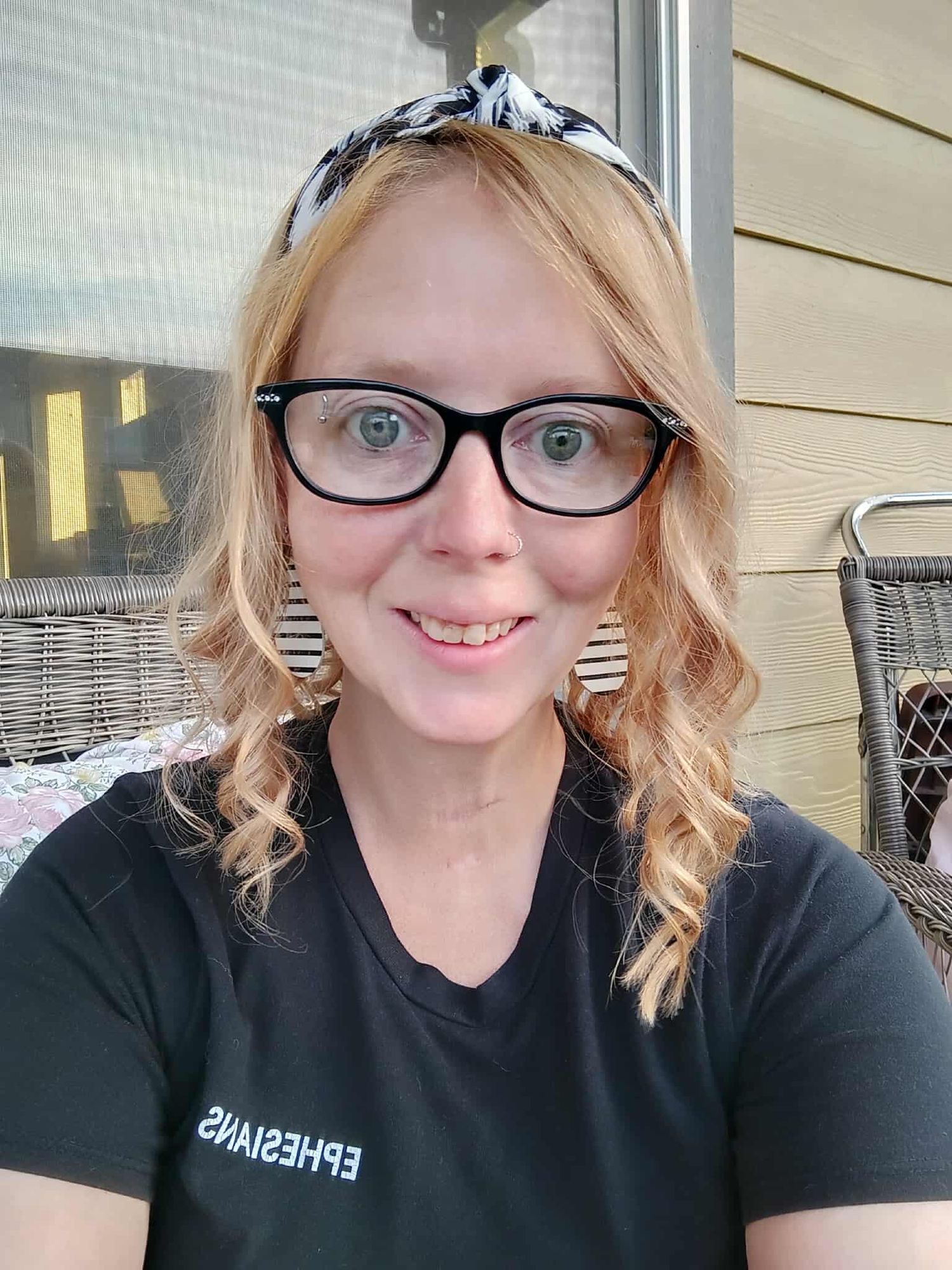 Woman wearing glasses and headband takes selfie outdoors on patio with wicker furniture.