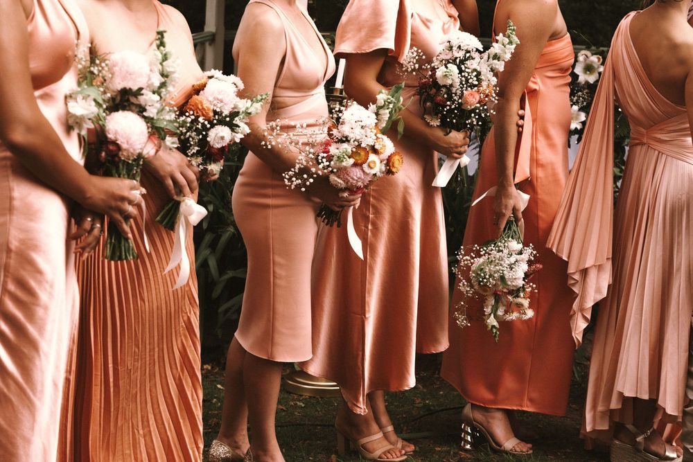 Bridesmaids stand in a line wearing various styles of coral and peach colored dresses while holding white floral bouquets.