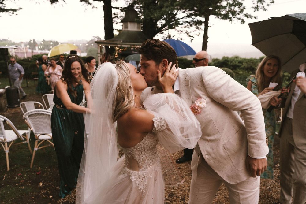 A joyful wedding celebration outdoors with guests gathering under tree cover.