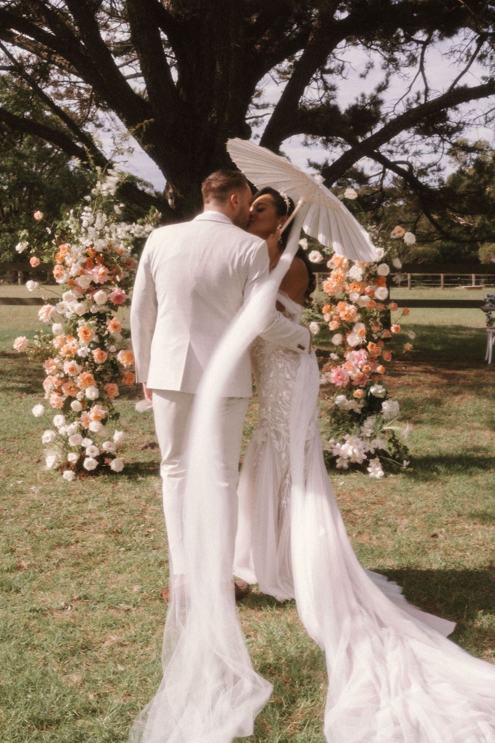 Romantic outdoor wedding ceremony with couple embracing under floral arch featuring peach and white roses in garden setting.