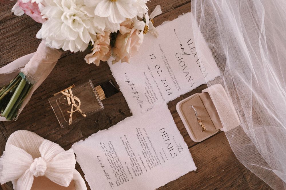 Wedding stationery and accessories including white invitations, sheer veil fabric, and soft pink flowers on a wooden surface.