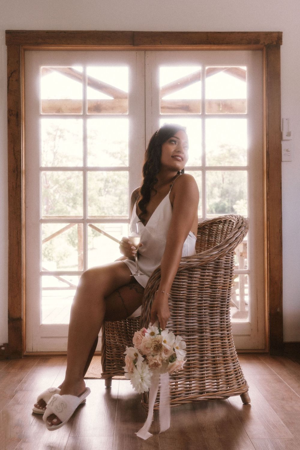A figure seated in a wicker chair by a bright window holds a delicate bouquet of light-colored flowers.