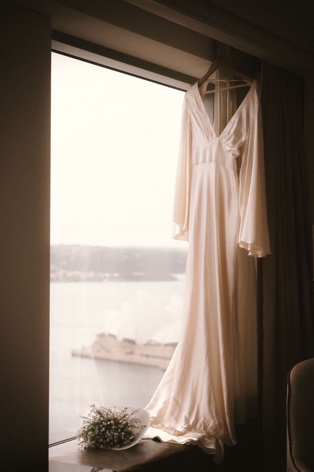 White wedding dress hangs by window in soft natural light creating ethereal atmosphere.