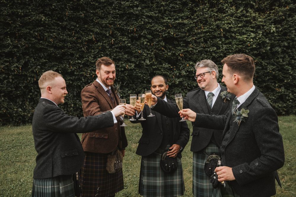 Groomsmen sharing laughs and drinks together during an outdoor wedding celebration in a garden setting.