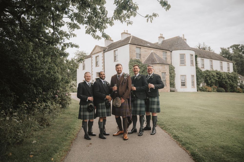 Wedding party enjoys outdoor photos in traditional Scottish kilts on green lawn in front of manor house at sunset.