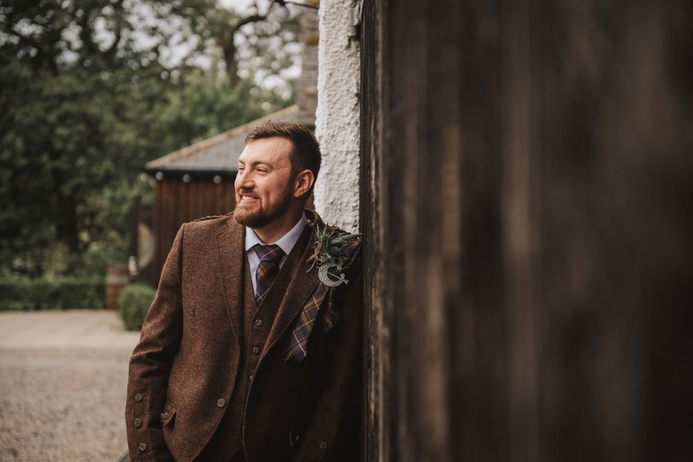 Person in brown tweed suit wearing a boutonniere leans against rustic wall in outdoor setting.