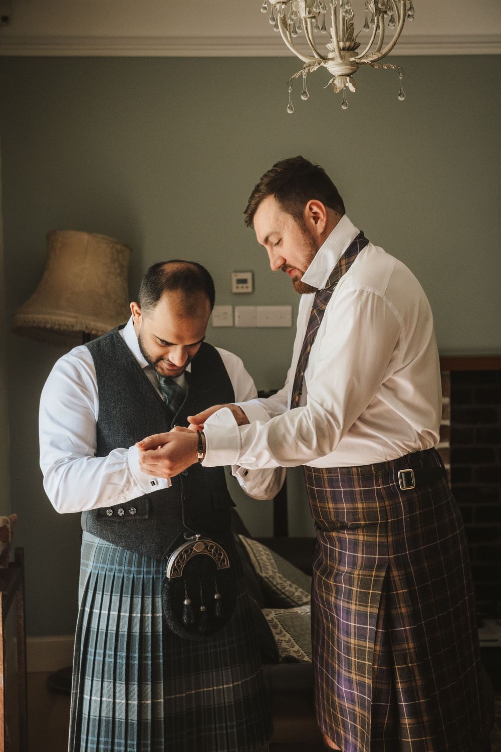 Two men in traditional Scottish dress help each other prepare formal attire before an event.