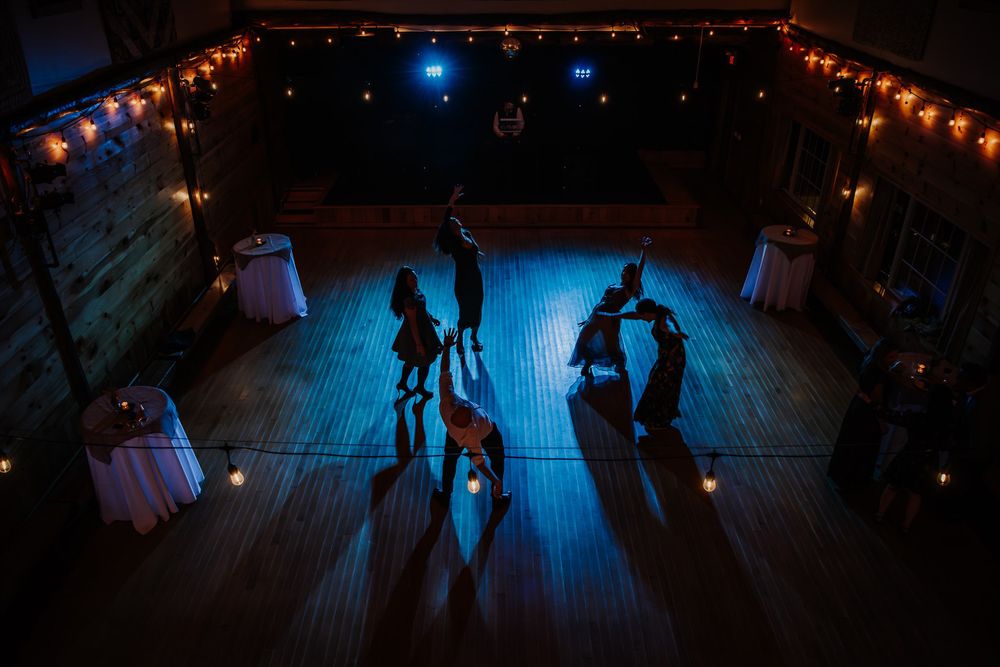 Silhouettes of people dancing under blue lighting and decorative string lights at a nighttime event.