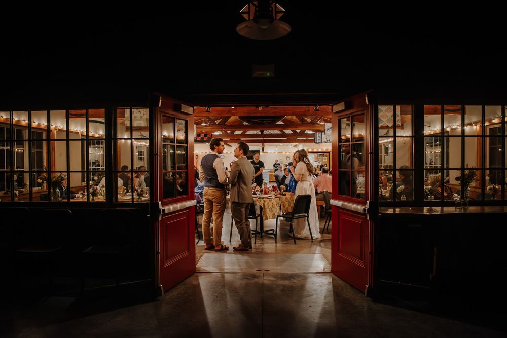 Wedding guests gather inside a warmly lit rustic restaurant venue with red barn-style doors at night.