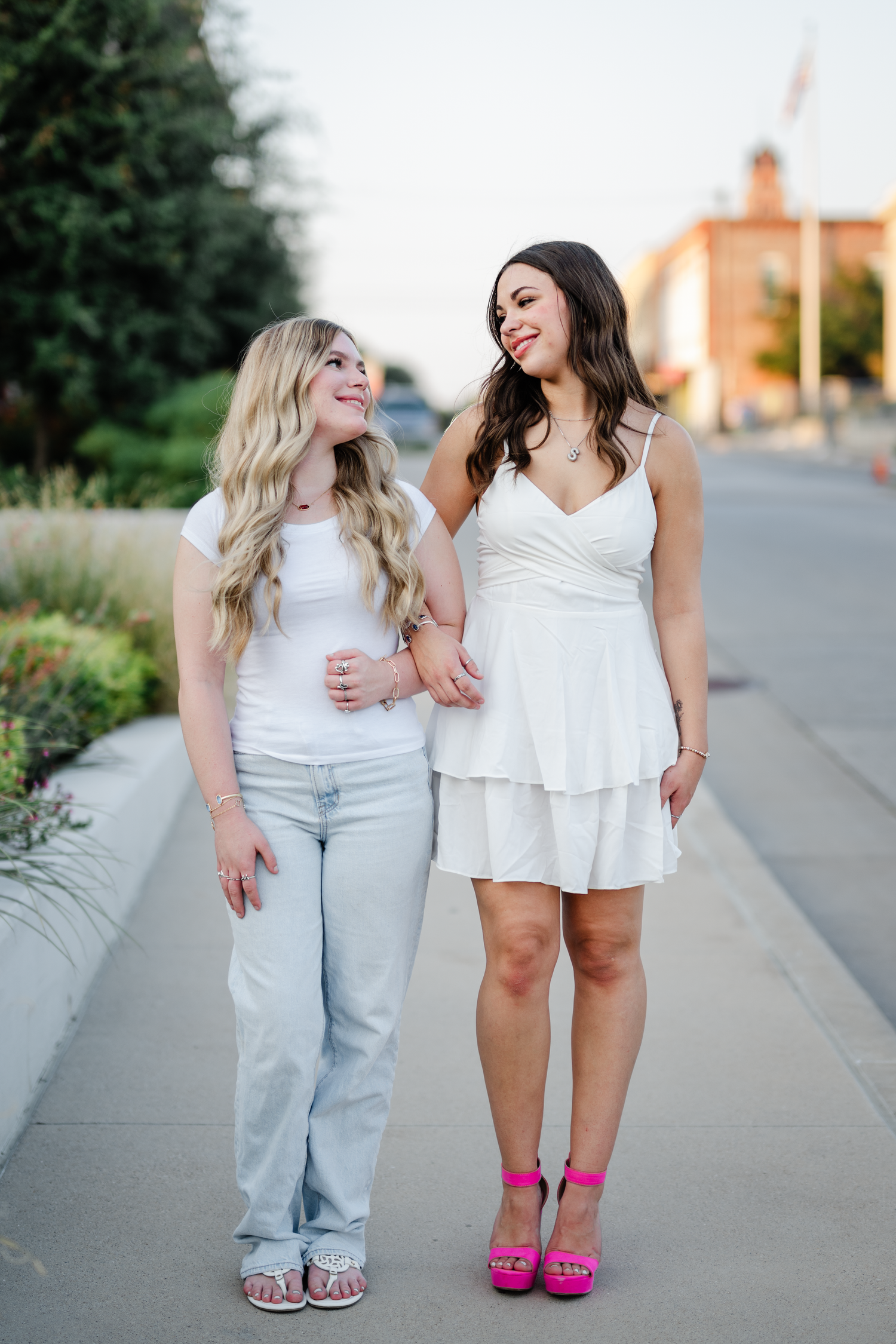 Two people in light colored outfits pose together and walk on a sidewalk, one wearing bright pink shoes.