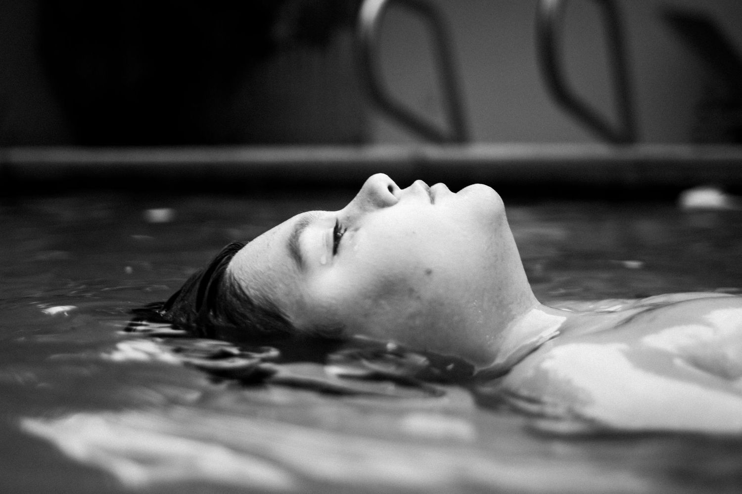 A person's face can be seen peacefully floating in dark reflective water with artistic black and white lighting.