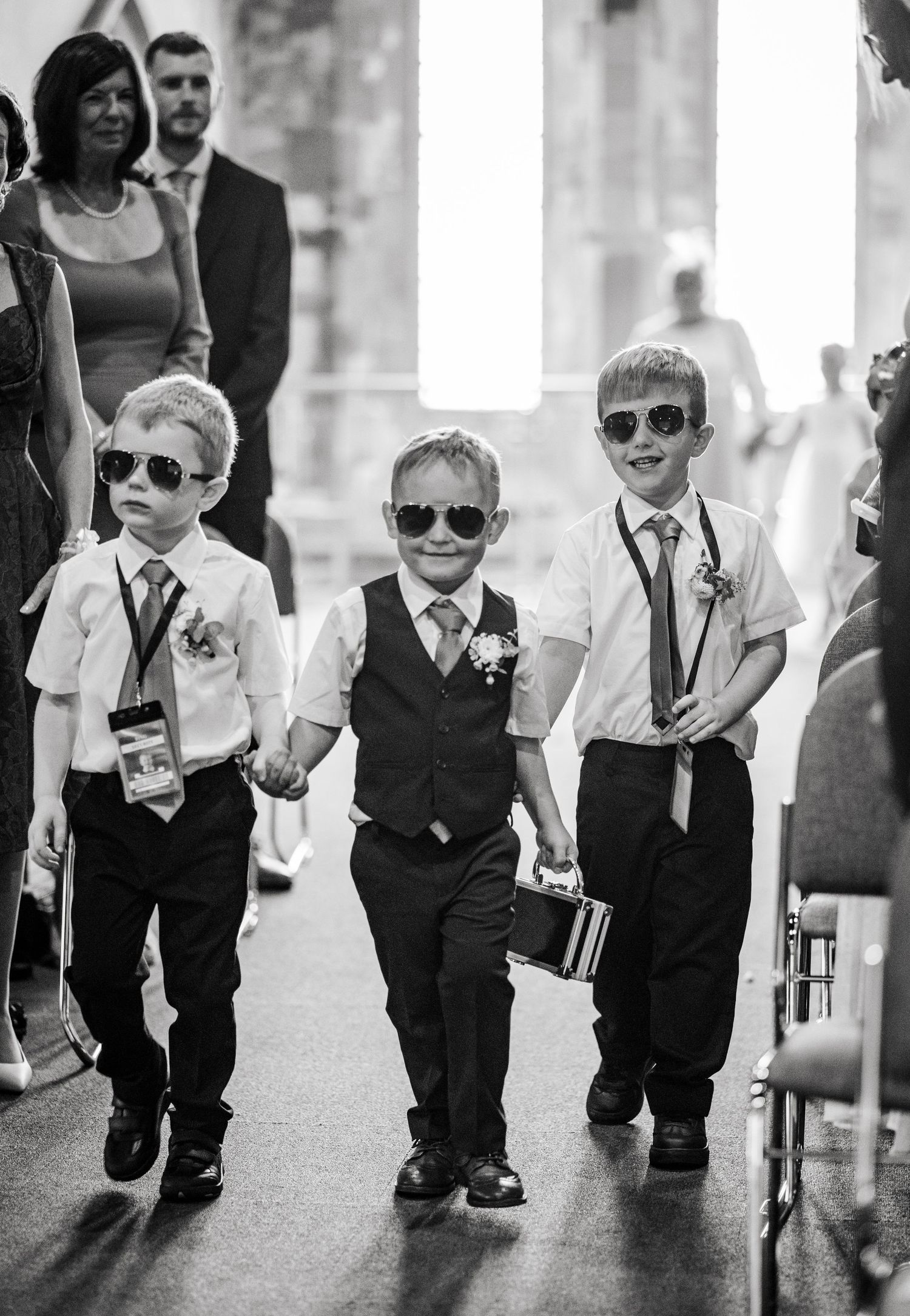 Wedding party featuring young ring bearers and page boys in matching sunglasses and formal attire walking down an aisle.