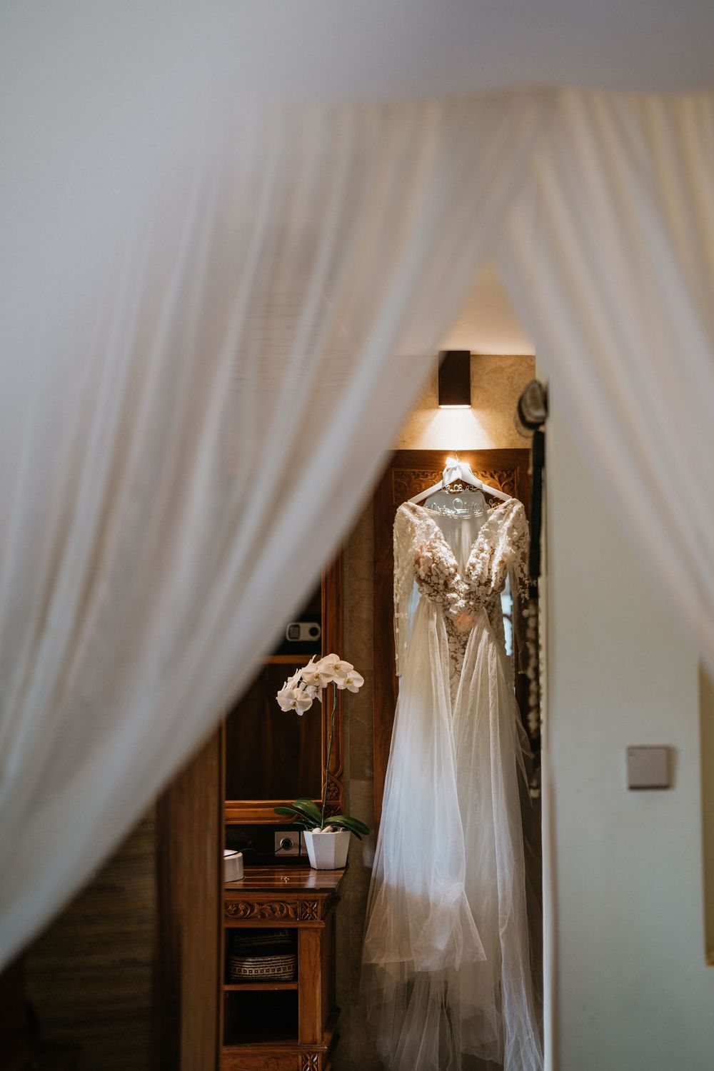 Elegant white wedding dress hanging against light curtains with delicate lace details.