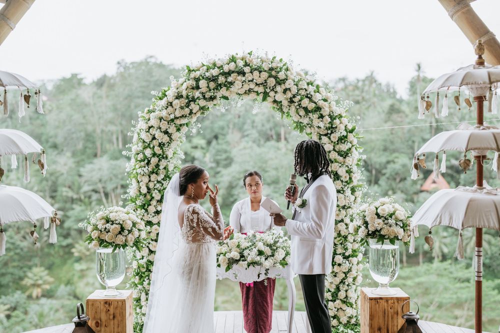 Wedding ceremony under white floral arch with tropical greenery backdrop.