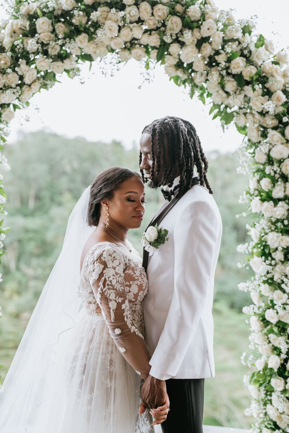 Romantic wedding moment under luxurious white rose arch with greenery.