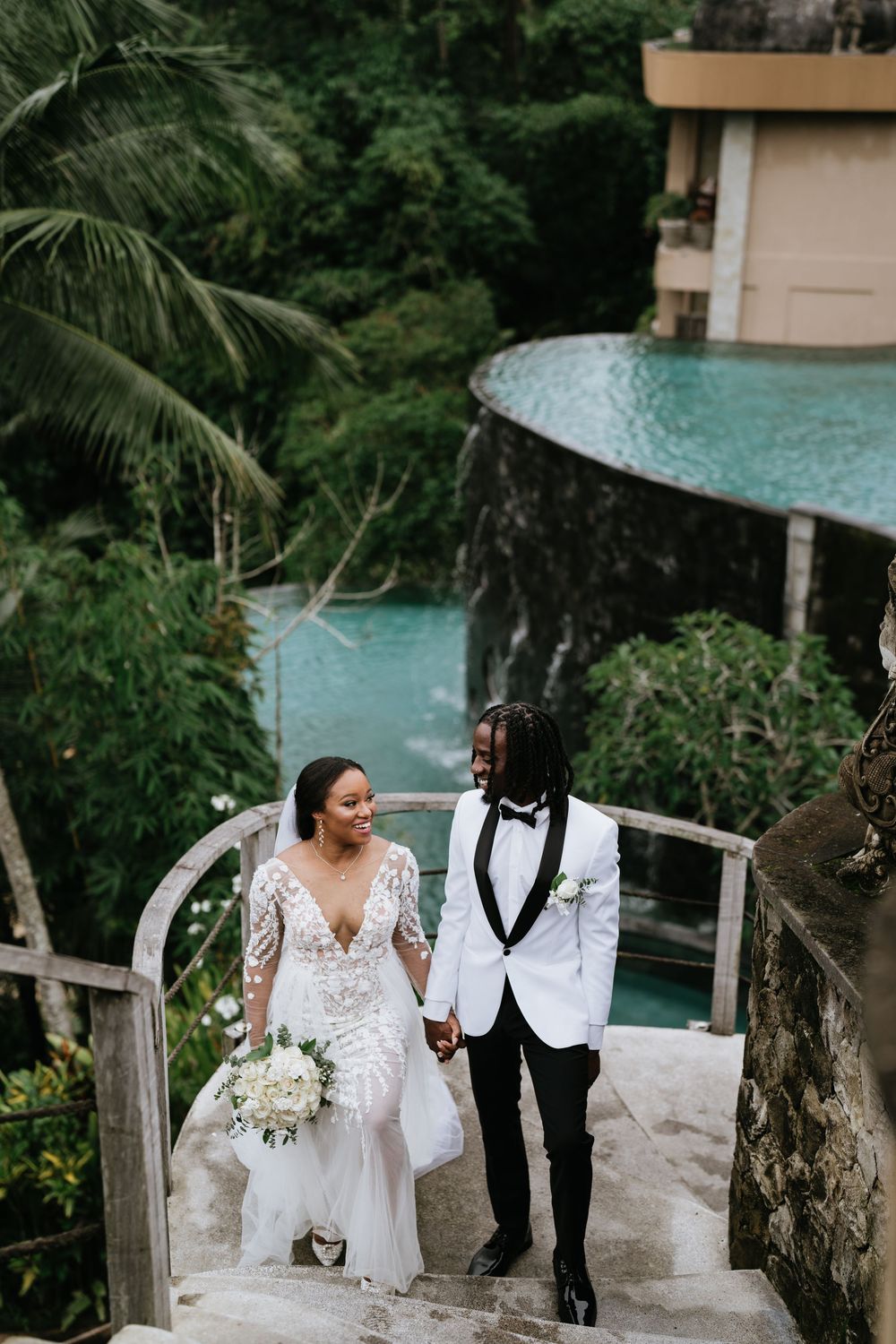 Newlyweds walking along infinity pool edge with tropical forest backdrop.