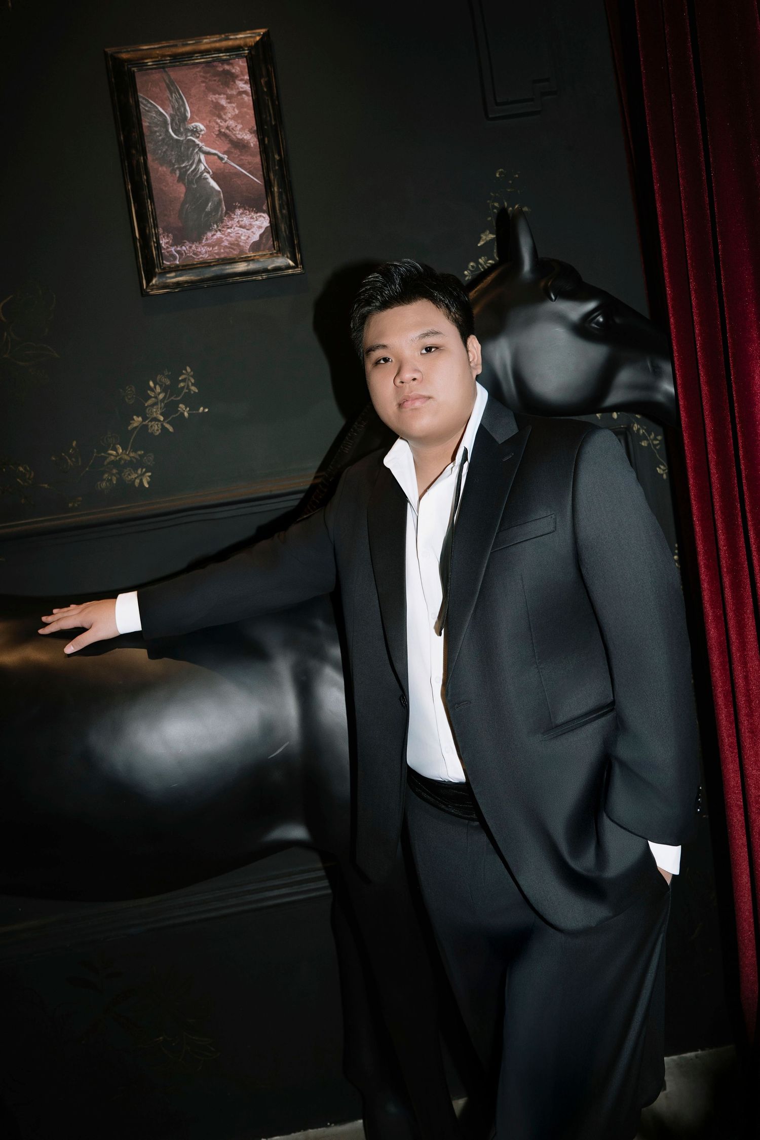 Asian groom in black suit posing beside large black animal sculpture in dark dramatic interior.