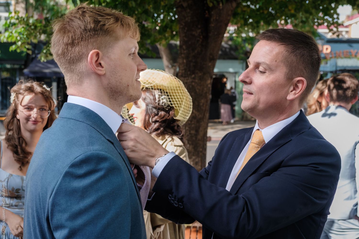 People in formal attire gathering at an outdoor social event with trees and guests mingling in the background.