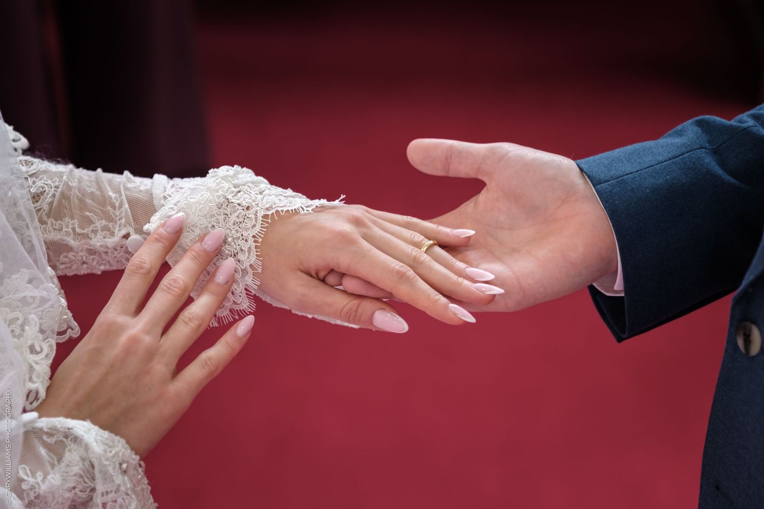 Close-up of hands exchanging wedding rings during marriage ceremony against red background.